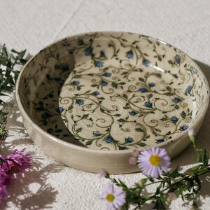 Könnte beinhalten: Eine runde Keramikschale mit cremefarbener Basis, verziert mit einem sich wiederholenden Muster aus dunkelgrünen Ranken und kleinen blauen Blüten. Die Schale hat eine glatte, glasierte Oberfläche und eine flache Form mit detaillierten botanischen Motiven.