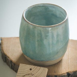 Könnte beinhalten: Eine handgefertigte, türkisfarbene Keramiktasse mit hellbeigem Boden, die auf einer Holzscheibe ruht. Ein kleines Holzschild mit einem Herz und Text befindet sich neben der Tasse. Die Tasse hat eine glatte, abgerundete Form.