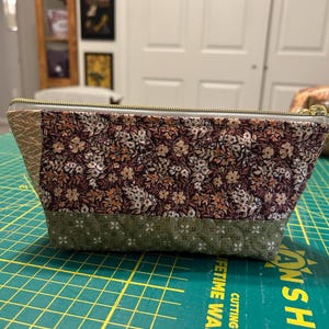 May include: A quilted fabric zipper pouch featuring a dark brown floral print on the upper section and a sage green patterned fabric on the lower section. The pouch has a gold-toned metal zipper closure and a side panel with a light tan leaf print.