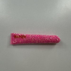 May include: A pink hair clip covered in small, sparkling rhinestones. The clip has a rectangular shape with a rounded end and a small, decorative bow near the opening. The clip is designed to hold hair in place.