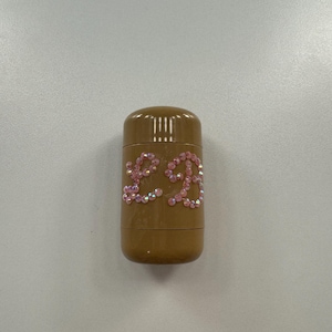 Op de afbeelding: Een bruinachtige, cilindervormige container met een geribbelde bovenkant. De container is versierd met de letters "£R" uit roze strass-steentjes. De container staat op een witte ondergrond.