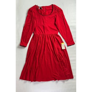 May include: A long-sleeved red dress with a round neckline and a flared skirt. The dress has black trim around the neckline and cuffs. A tag is attached to the dress. The dress is made of a soft, flowing fabric.