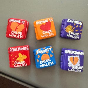 May include: Six miniature, colorful book-shaped objects. Each has a different title and design, including hearts, butterflies, and abstract shapes. The author's name, Chloe Walsh, is written on each.
