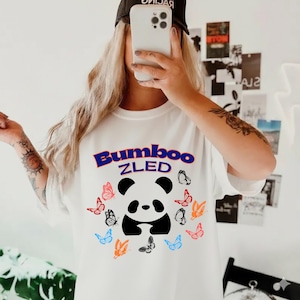 May include: White t-shirt featuring a panda bear design encircled by colourful butterflies. The text "Bumboo ZLED" is above the panda in blue and purple lettering. A casual style t-shirt.
