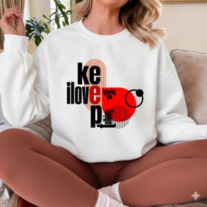 May include: White sweatshirt featuring the text "keep ignoring you" in black, red, and white. The design includes a red circle and black lines. A casual style sweatshirt.