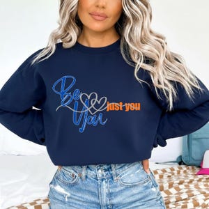 Op de afbeelding: Marineblauwe sweatshirt met de woorden "Be You" in een scriptlettertype, met een hartontwerp en de woorden "just you" in oranje. De sweatshirt heeft een ronde hals.