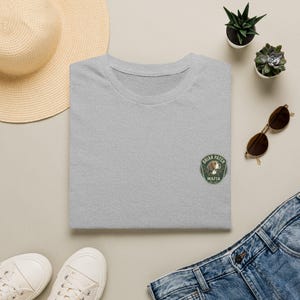 Pode incluir: Uma t-shirt cinzenta mesclada com um emblema circular bordado no peito. O emblema é verde-escuro com uma borda branca e apresenta uma ilustração de um cão castanho e branco com o texto "BRIAR PATCH MAFIA" e "EST 2017".