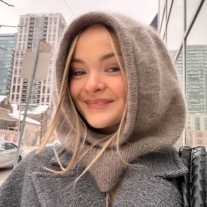 Könnte beinhalten: Ein hellbraunes Strick-Hoodie- und Schal-Set. Die Kapuze ist hochgezogen und umrahmt das Gesicht. Die Person trägt einen grauen Mantel. Der Hintergrund zeigt eine Stadtansicht mit Gebäuden und einem bewölkten Himmel.