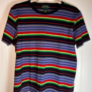 Puede incluir: Camiseta de manga corta a rayas en tonos azules, negros, rojos, amarillos y verdes. La camiseta tiene cuello redondo y el nombre de la marca "Lauren" es visible en la etiqueta. Cuelga de una percha blanca.