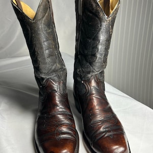 Op de afbeelding: Een paar versleten, bruine leren cowboylaarzen. De laarzen hebben een vierkante neus en decoratieve stiksels op de wreef. De schachten zijn donkerbruin en zwart, met een treklus aan de bovenkant.