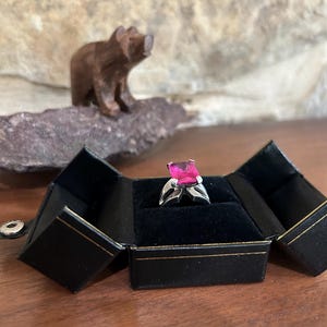 May include: A silver ring with a large, square, pink gemstone is displayed in an open black jewelry box with gold trim. A small, carved wooden bear sits in the background.