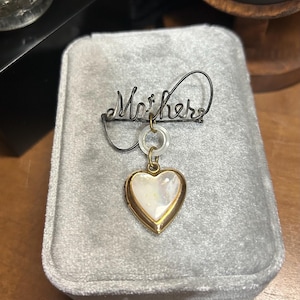 May include: A vintage brooch with the word "Mother" in cursive script. A gold-toned heart pendant with a pearlescent center hangs below. The brooch is set on a gray velvet surface.