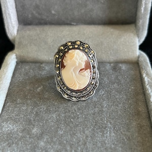 Könnte beinhalten: Ovale Cameo-Ring mit Profil zweier Frauen in Creme- und Brauntönen. Der Ring hat eine silberfarbene Fassung mit einem dekorativen Rand aus kleinen, runden Akzenten. Der Ring wird in einer grauen Samt-Schmuckschatulle präsentiert.