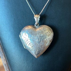 May include: A silver-toned heart-shaped locket pendant suspended from a delicate box chain. The locket features an intricate, engraved floral pattern across its front surface, with a polished bail connecting it to the thin metal necklace chain.