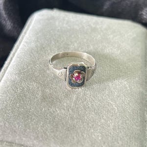 May include: A silver-toned ring featuring a rectangular face with a textured dark blue inlay. At the center of the inlay is a small, five-petaled flower motif holding a single, round, vibrant pink gemstone. The band has engraved detailing.