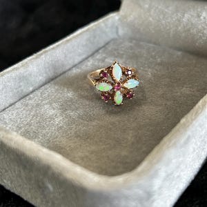 Op de afbeelding: Een gouden ring met een centraal clusterontwerp van vier marquise-geslepen opalen in een kruisvorm, afgewisseld met vier ronde, donkerroze edelstenen. De zetting is vervaardigd uit geelgoud en heeft een gedetailleerd metalen frame.