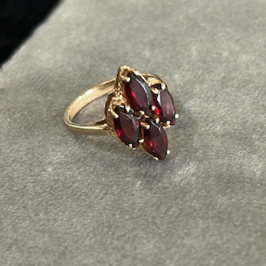 May include: A gold ring featuring a cluster of five marquise-cut deep red garnet gemstones arranged in a vertical, pointed formation. The metal band is smooth and polished, supporting the faceted stones which are set in a secure metal mounting.