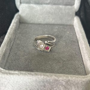 Può includere: Un anello bypass color argento con due incastonature quadrate affiancate. Una incastonatura contiene una pietra trasparente a taglio rotondo, mentre l'altra contiene una vivace pietra rotonda color rosa-rosso. La fascia ha una finitura metallica liscia.
