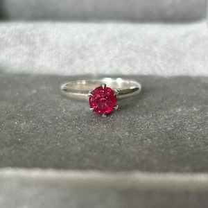 May include: A silver ring with a vibrant red gemstone. The gemstone is round and set in a six-prong setting. The ring is displayed on a gray surface.