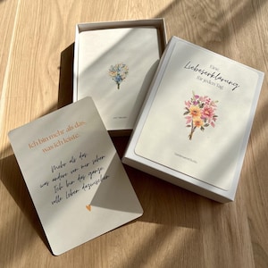 Könnte beinhalten: Eine weiße Schachtel mit einem Kartenset, das florale Illustrationen und deutschen Text enthält. Eine Karte zeigt einen Blumenstrauß und den Titel Eine Liebeserklärung für jeden Tag. Eine weitere Karte trägt den Text Ich bin mehr als das, was ich leiste.