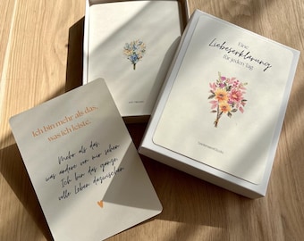 Könnte beinhalten: Eine weiße Schachtel mit einem Kartenset, das florale Illustrationen und deutschen Text enthält. Eine Karte zeigt einen Blumenstrauß und den Titel Eine Liebeserklärung für jeden Tag. Eine weitere Karte trägt den Text Ich bin mehr als das, was ich leiste.