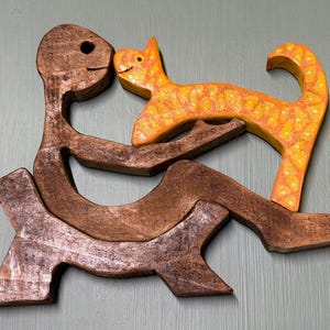 May include: A dark brown wooden puzzle piece shaped like a person sitting down, holding a bright orange cat puzzle piece with yellow spots. The two interlocking wooden figures form a cohesive shape with smooth edges and a natural wood grain finish.