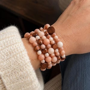 Può includere: Un bracciale a più fili caratterizzato da una combinazione di perline rotonde e lisce nei toni del pesca e della crema, alternate a perline in legno marrone scuro di varie forme, tra cui dischi rotondi, cilindrici e piatti su cordino elastico.