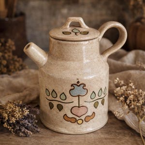 Può includere: Una teiera in ceramica beige con coperchio e manico. La teiera è decorata con un motivo floreale blu, verde, rosa e giallo. La teiera è appoggiata su una superficie di legno.