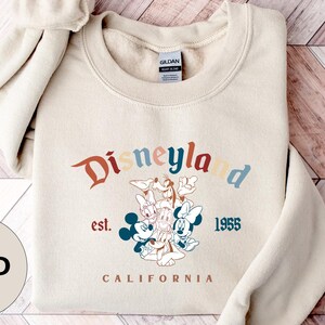 Puede incluir: Sudadera de Disneylandia color arena con cuello redondo. La parte delantera presenta el logotipo de Disneylandia en colores retro, el texto "est. 1955 CALIFORNIA" y un gr&aacute;fico de Mickey y Minnie Mouse, Donald Duck y Goofy.