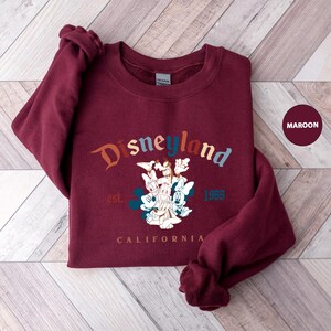 Puede incluir: Sudadera granate con la palabra "Disneyland" en una fuente arqueada y colorida. Debajo del texto hay un gr&aacute;fico de personajes de Disney, con el texto "est. 1955" y "CALIFORNIA". La sudadera es de color granate liso.