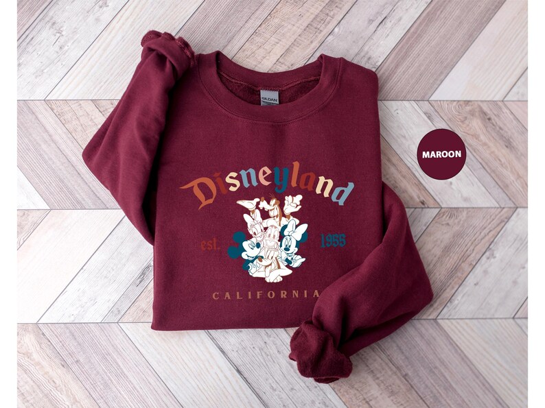 Puede incluir: Sudadera granate con la palabra "Disneyland" en una fuente arqueada y colorida. Debajo del texto hay un gr&aacute;fico de personajes de Disney, con el texto "est. 1955" y "CALIFORNIA". La sudadera es de color granate liso.