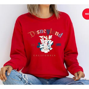 Puede incluir: Una sudadera roja con el logotipo de Disneyland en un dise&ntilde;o multicolor. El gr&aacute;fico incluye a Mickey Mouse, Minnie Mouse, el Pato Donald y Goofy. Tambi&eacute;n se ve el texto "est. 1955 CALIFORNIA".