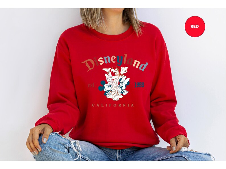 Puede incluir: Una sudadera roja con el logotipo de Disneyland en un dise&ntilde;o multicolor. El gr&aacute;fico incluye a Mickey Mouse, Minnie Mouse, el Pato Donald y Goofy. Tambi&eacute;n se ve el texto "est. 1955 CALIFORNIA".
