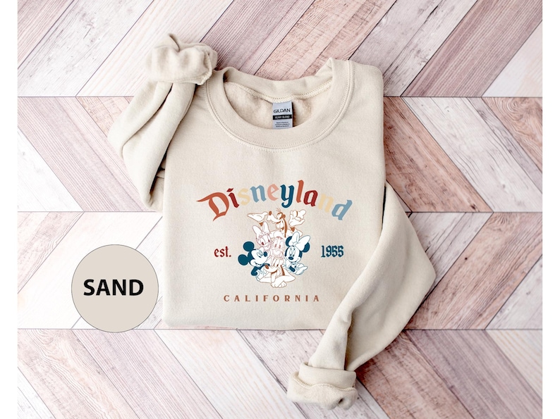Puede incluir: Sudadera de Disneylandia color arena con cuello redondo. La parte delantera presenta el logotipo de Disneylandia en colores retro, el texto "est. 1955 CALIFORNIA" y un gr&aacute;fico de Mickey y Minnie Mouse, Donald Duck y Goofy.