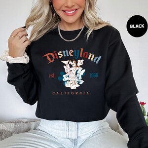 Puede incluir: Sudadera negra de Disneylandia con un gr&aacute;fico colorido con Mickey Mouse, Minnie Mouse y otros personajes. El texto "Disneyland" est&aacute; arqueado en la parte superior, con "est. 1955" y "CALIFORNIA" debajo del gr&aacute;fico. Un c&iacute;rculo negro con la palabra "BLACK" est&aacute; en la parte superior derecha.