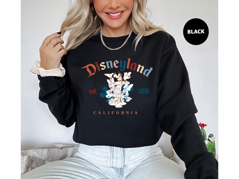 Puede incluir: Sudadera negra de Disneylandia con un gr&aacute;fico colorido con Mickey Mouse, Minnie Mouse y otros personajes. El texto "Disneyland" est&aacute; arqueado en la parte superior, con "est. 1955" y "CALIFORNIA" debajo del gr&aacute;fico. Un c&iacute;rculo negro con la palabra "BLACK" est&aacute; en la parte superior derecha.