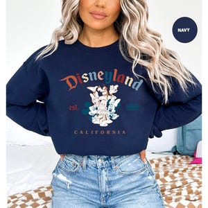 Puede incluir: Sudadera azul marino de Disneyland con un gr&aacute;fico colorido. El dise&ntilde;o presenta el logotipo de Disneyland, Mickey Mouse y otros personajes. El texto "Disneyland" est&aacute; arqueado en la parte superior, con "est. 1955" y "California" debajo.