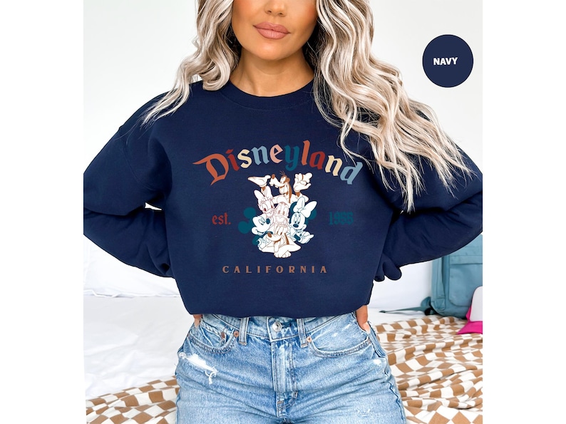 Puede incluir: Sudadera azul marino de Disneyland con un gr&aacute;fico colorido. El dise&ntilde;o presenta el logotipo de Disneyland, Mickey Mouse y otros personajes. El texto "Disneyland" est&aacute; arqueado en la parte superior, con "est. 1955" y "California" debajo.
