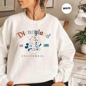 Puede incluir: Sudadera blanca de cuello redondo con el logotipo de Disneyland, con Mickey Mouse, Minnie Mouse, Donald Duck y Daisy Duck. El dise&ntilde;o incluye el texto "Disneyland est. 1955 California" en una fuente y esquema de color retro.