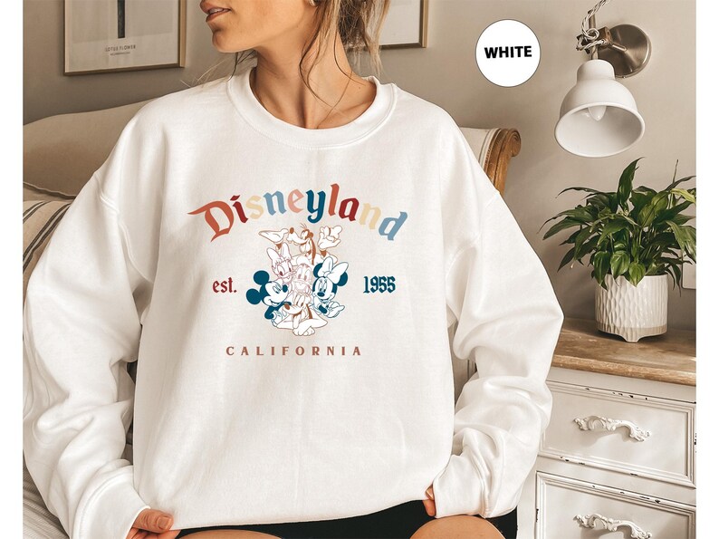 Puede incluir: Sudadera blanca de cuello redondo con el logotipo de Disneyland, con Mickey Mouse, Minnie Mouse, Donald Duck y Daisy Duck. El dise&ntilde;o incluye el texto "Disneyland est. 1955 California" en una fuente y esquema de color retro.