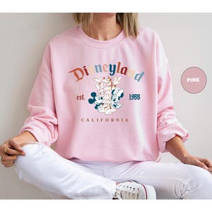 Puede incluir: Sudadera rosa con el logotipo de Disneyland en colores arco&iacute;ris. El dise&ntilde;o incluye a Mickey Mouse, Minnie Mouse, Donald Duck y Goofy. Tambi&eacute;n est&aacute; impreso el texto "est. 1955 CALIFORNIA".