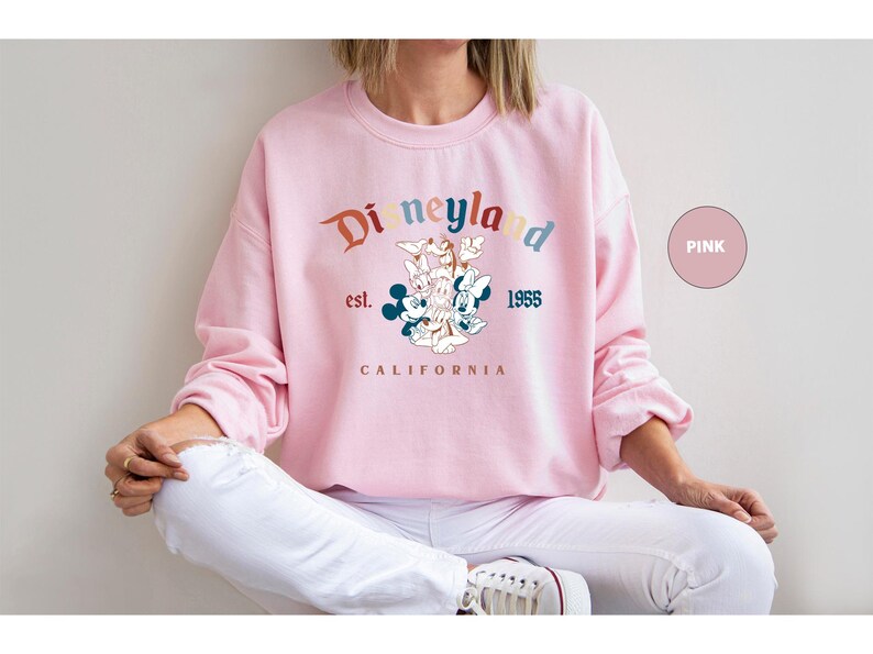 Puede incluir: Sudadera rosa con el logotipo de Disneyland en colores arco&iacute;ris. El dise&ntilde;o incluye a Mickey Mouse, Minnie Mouse, Donald Duck y Goofy. Tambi&eacute;n est&aacute; impreso el texto "est. 1955 CALIFORNIA".
