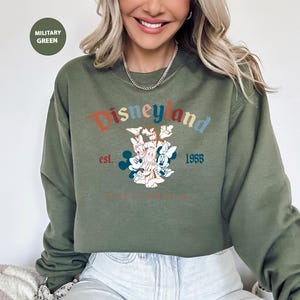 Puede incluir: Sudadera verde militar con el logotipo de Disneyland en texto colorido y un gr&aacute;fico de Mickey y Minnie Mouse, Donald Duck y Daisy Duck. La sudadera tambi&eacute;n tiene el texto "est. 1955 CALIFORNIA".