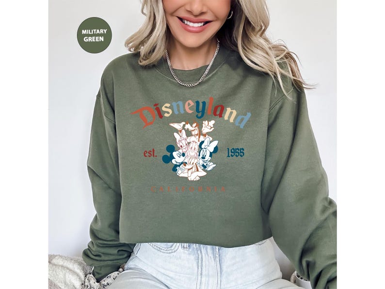 Puede incluir: Sudadera verde militar con el logotipo de Disneyland en texto colorido y un gr&aacute;fico de Mickey y Minnie Mouse, Donald Duck y Daisy Duck. La sudadera tambi&eacute;n tiene el texto "est. 1955 CALIFORNIA".