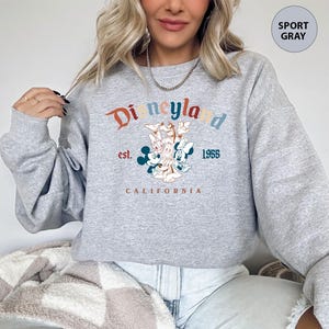Puede incluir: Sudadera gris deportiva con un gr&aacute;fico de Disneyland. El dise&ntilde;o presenta el logotipo de Disneyland en naranja, verde azulado y azul, con Mickey y Minnie Mouse. Tambi&eacute;n se incluye el texto "est. 1955 CALIFORNIA".