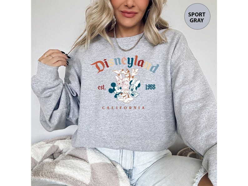 Puede incluir: Sudadera gris deportiva con un gr&aacute;fico de Disneyland. El dise&ntilde;o presenta el logotipo de Disneyland en naranja, verde azulado y azul, con Mickey y Minnie Mouse. Tambi&eacute;n se incluye el texto "est. 1955 CALIFORNIA".
