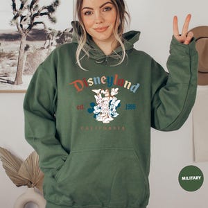 Puede incluir: Una sudadera con capucha verde oliva con un bolsillo delantero y la palabra "Disneyland" en una fuente colorida de estilo vintage. La sudadera presenta un gr&aacute;fico de Mickey Mouse, Minnie Mouse y Donald Duck, con "est. 1955 CALIFORNIA" impreso debajo.