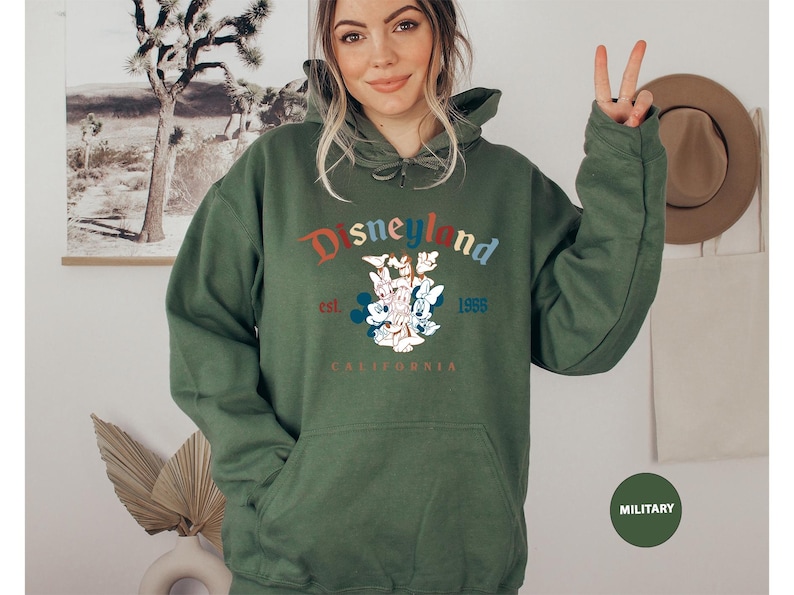 Puede incluir: Una sudadera con capucha verde oliva con un bolsillo delantero y la palabra "Disneyland" en una fuente colorida de estilo vintage. La sudadera presenta un gr&aacute;fico de Mickey Mouse, Minnie Mouse y Donald Duck, con "est. 1955 CALIFORNIA" impreso debajo.