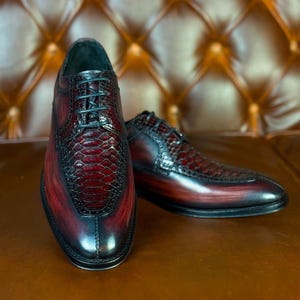 May include: A pair of leather dress shoes with a gradient burgundy and black finish. The toe and vamp feature a textured, reptile-like pattern. Black laces and a black sole complete the formal look.