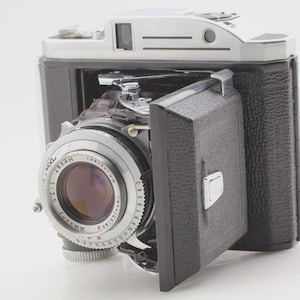 May include: A vintage folding camera with a black textured body and silver metal top plate. The lens assembly features a Konishiroku Hexar 75mm f/3.5 lens with MXL markings. The front door is hinged and black, revealing the lens and shutter mechanism.